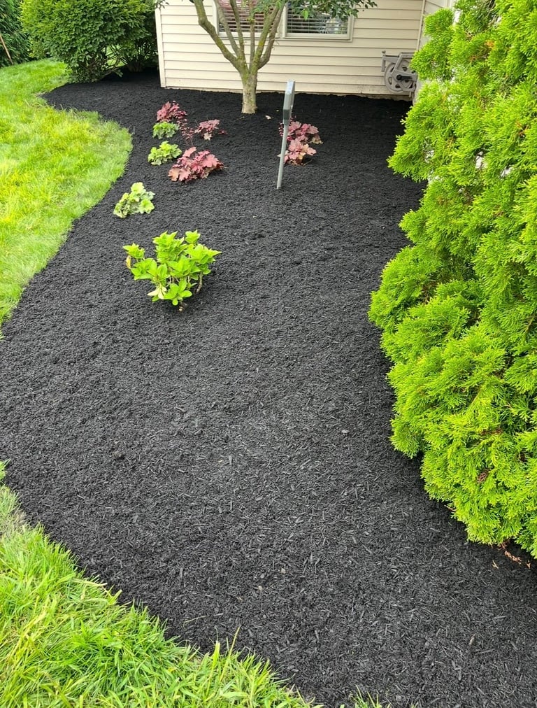 Landscaped garden bed with dark mulch, pink and green flowering plants, and bright green shrubs surrounding a residential home