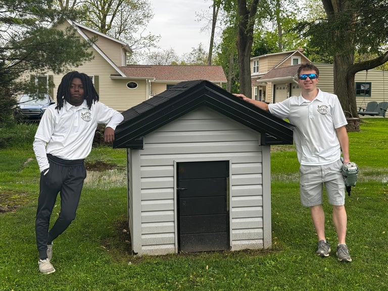 Two workers in white shirts posing with a small white dog house with black roof in a residential yard